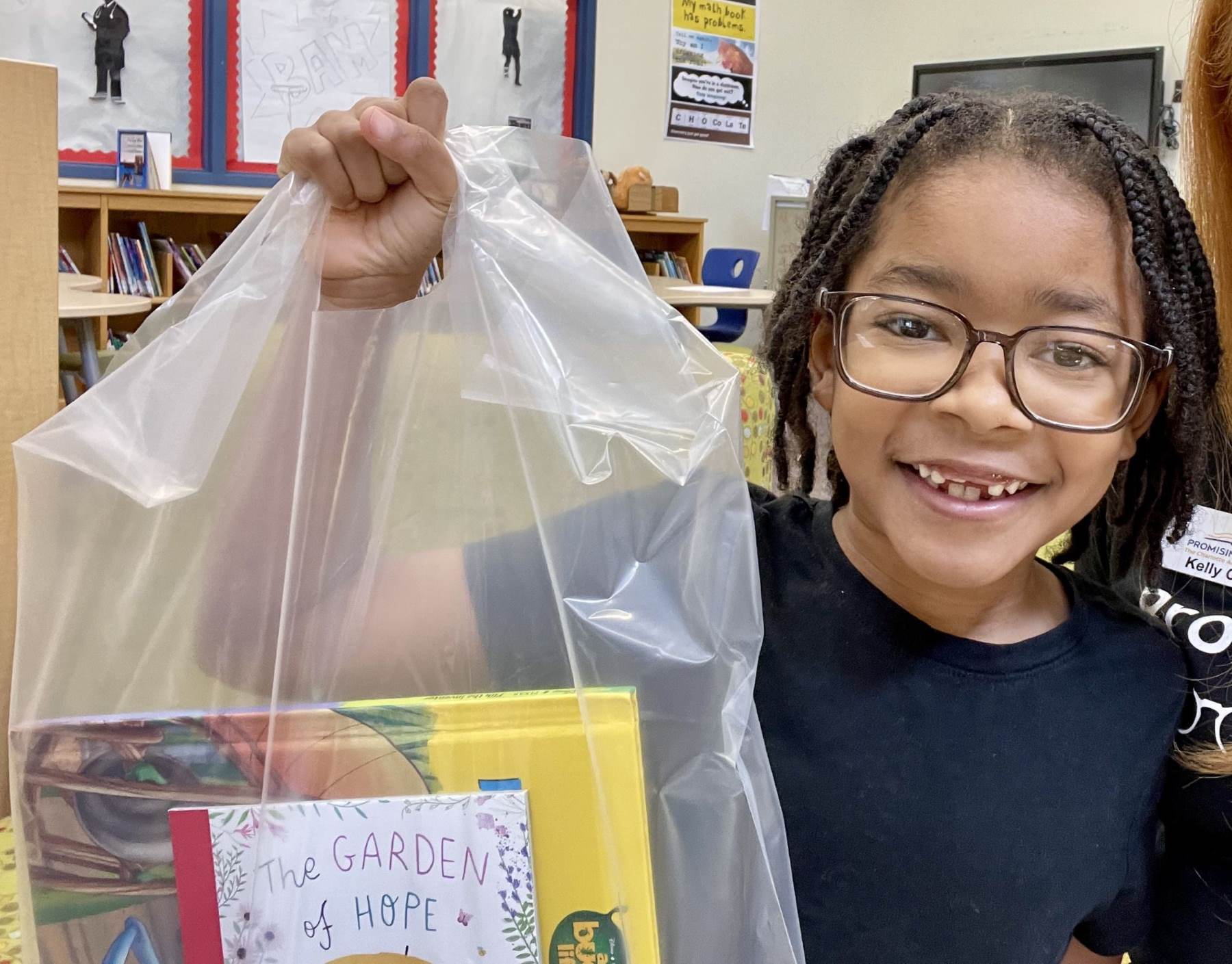Black boy with glasses and and a navy t-shirt holding up a plastic bag with books in it. He is looking at the camera smiling.