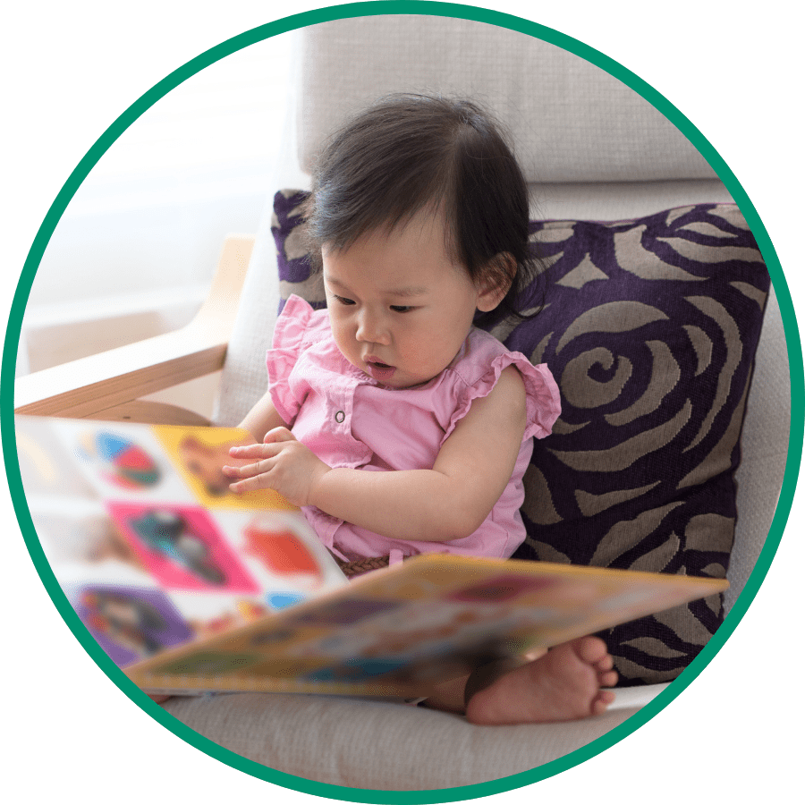 A little girl picking holding a book in her hands and picking out another one from a book shelf. She is looking at the book she's picking up and starting to smile.