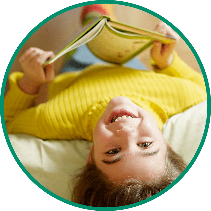 White girl sitting upside down over a couch with a book in her hands smiling at the camera.
