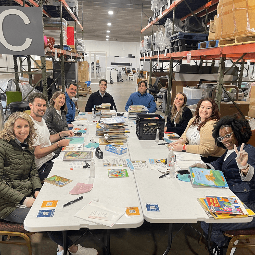 Group of 9 diverse people sitting around a table with books and cleaning supplies. They are all looking at the camera smiling. 
