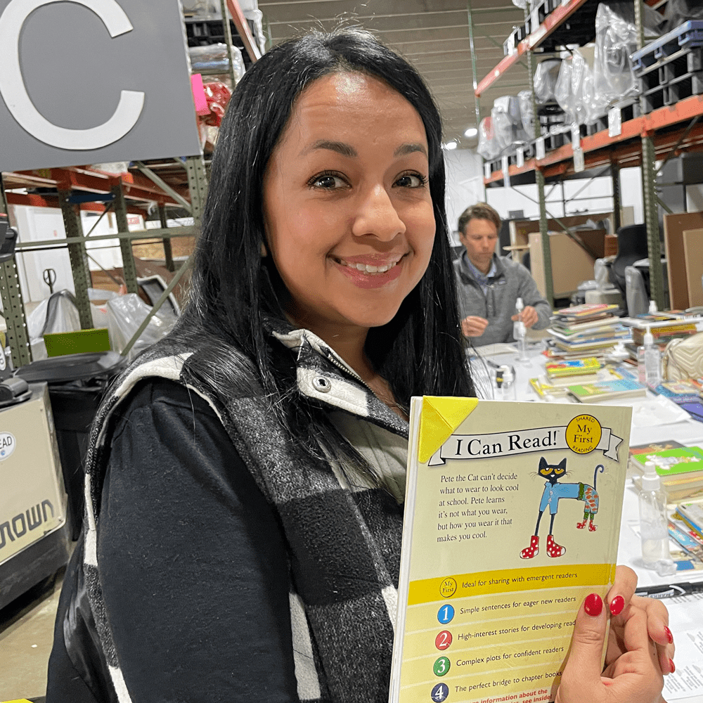 South Asian woman holding up a book smiling at the camera while volunteering at the Promising Pages warehouse. 