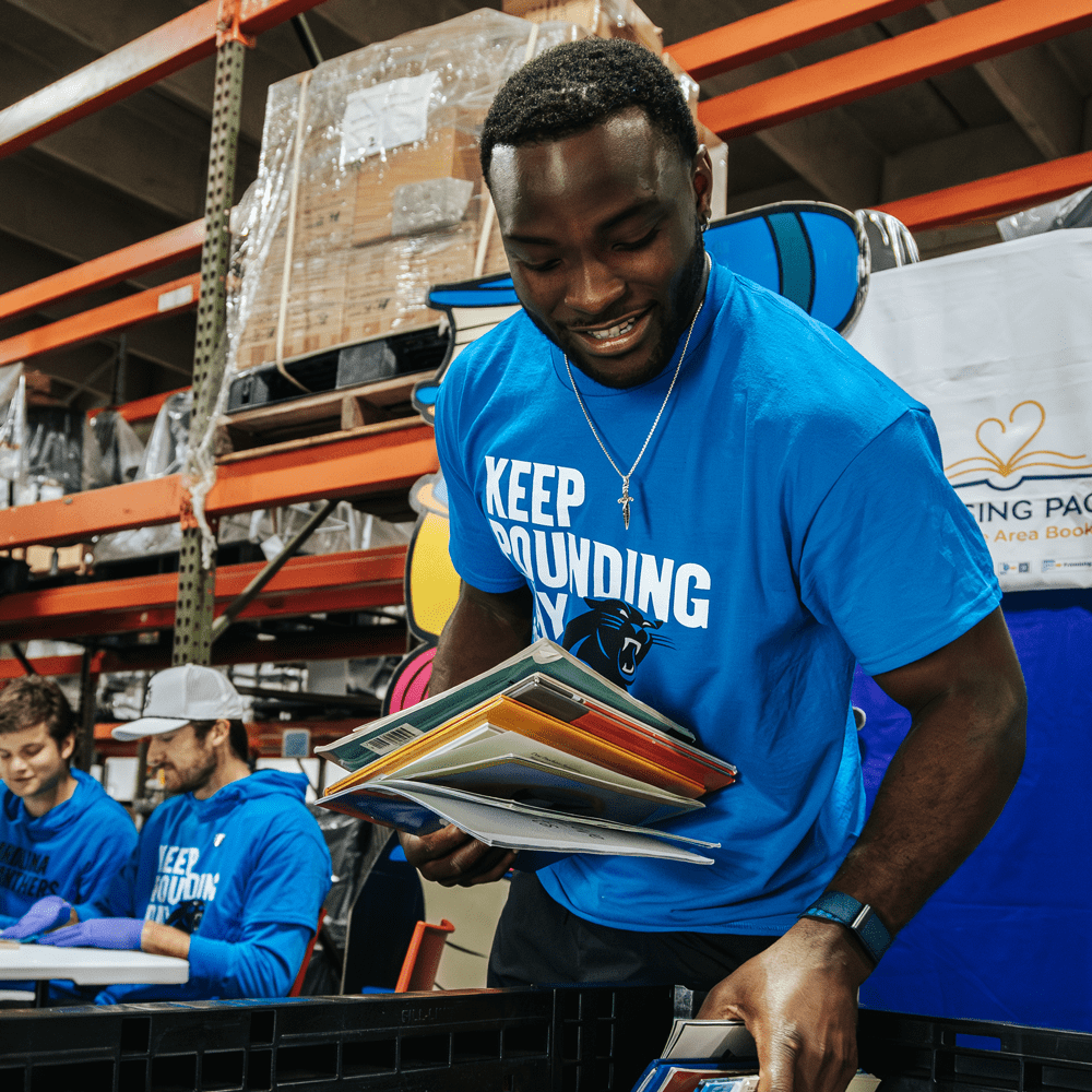 Man looking holding a stack of books while looking at a bin. Behind him are two other people sitting at a table cleaning books. All have Carolina Panthers shirts that say "Keep Pounding Day".
