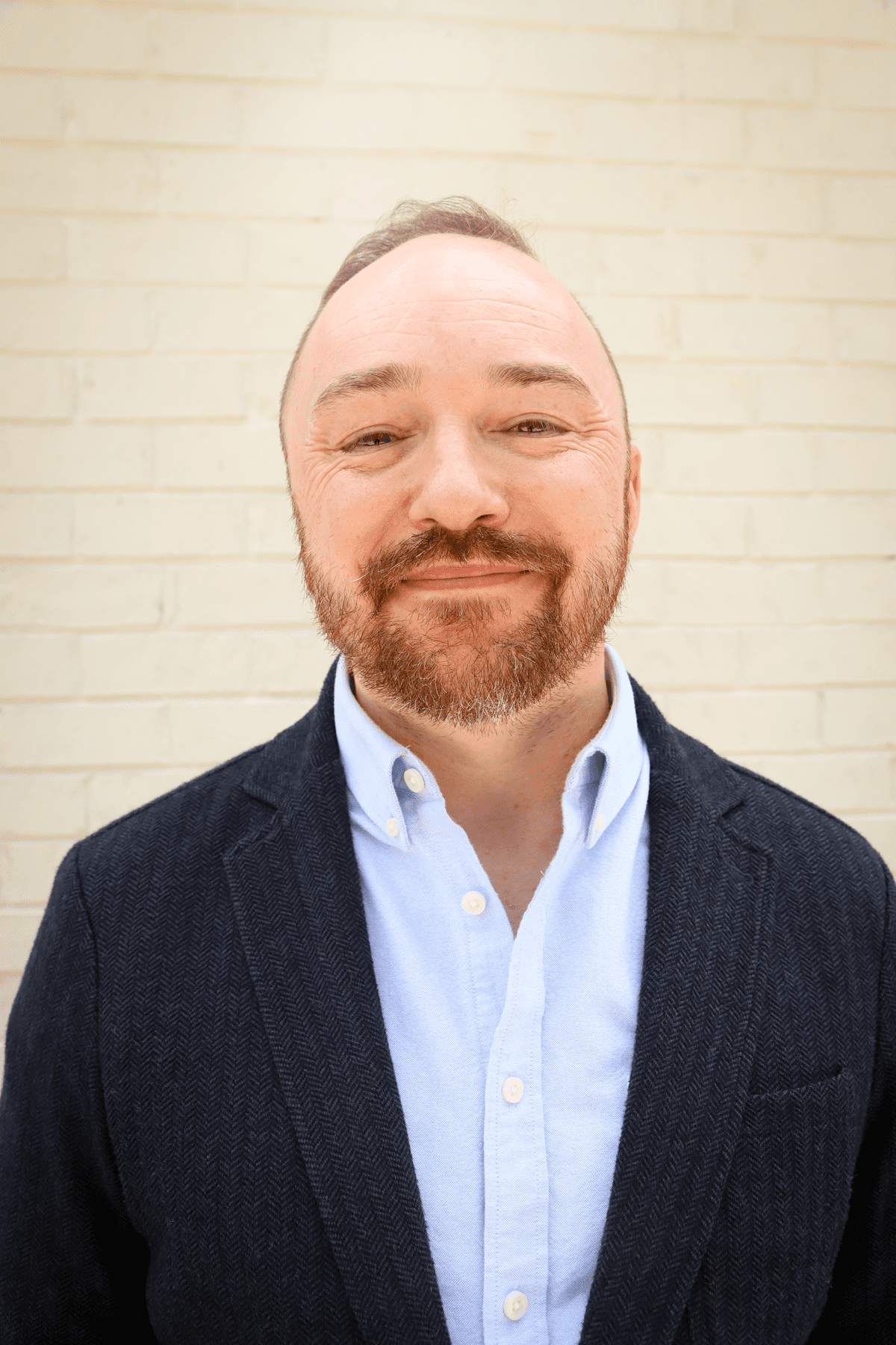 A white man with brown hair and facial hair wearing a black blazer with a blue button up. He is looking at the camera smiling.